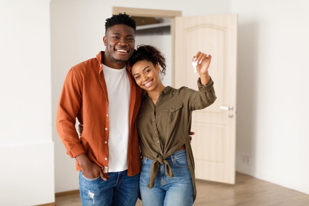 Homebuying as an Unmarried Couple: What to Consider. Real Estate. Cheerful African American Spouses Showing New House Key Smiling To Camera, Embracing Standing Among Moving Cardboard Boxes At Home. Relocation, Apartment Ownership And Family Housing