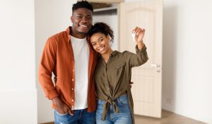 Homebuying as an Unmarried Couple: What to Consider. Real Estate. Cheerful African American Spouses Showing New House Key Smiling To Camera, Embracing Standing Among Moving Cardboard Boxes At Home. Relocation, Apartment Ownership And Family Housing
